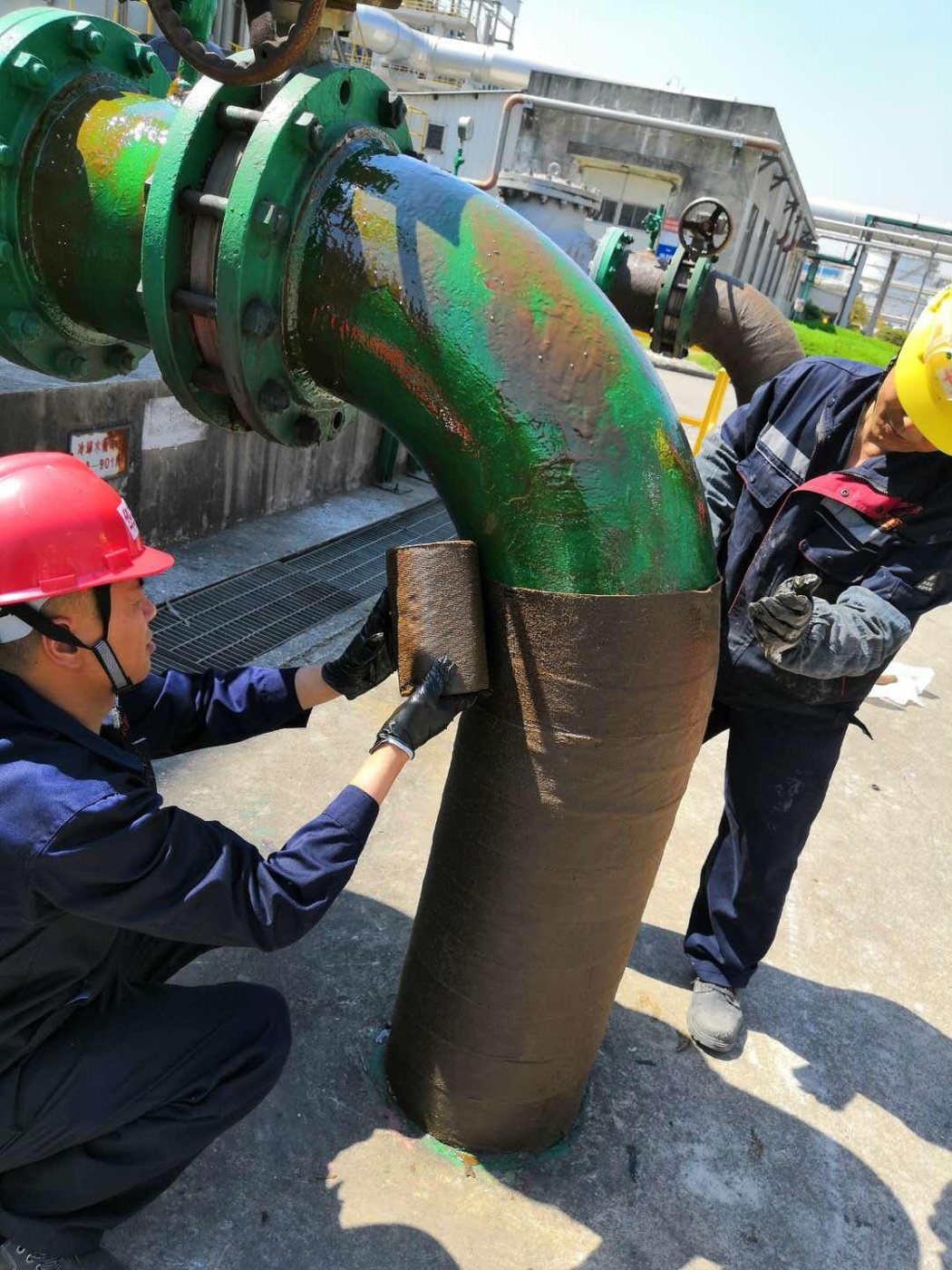 Two applicators wrapping a pipe bend with petrolatum tape on an operating plant